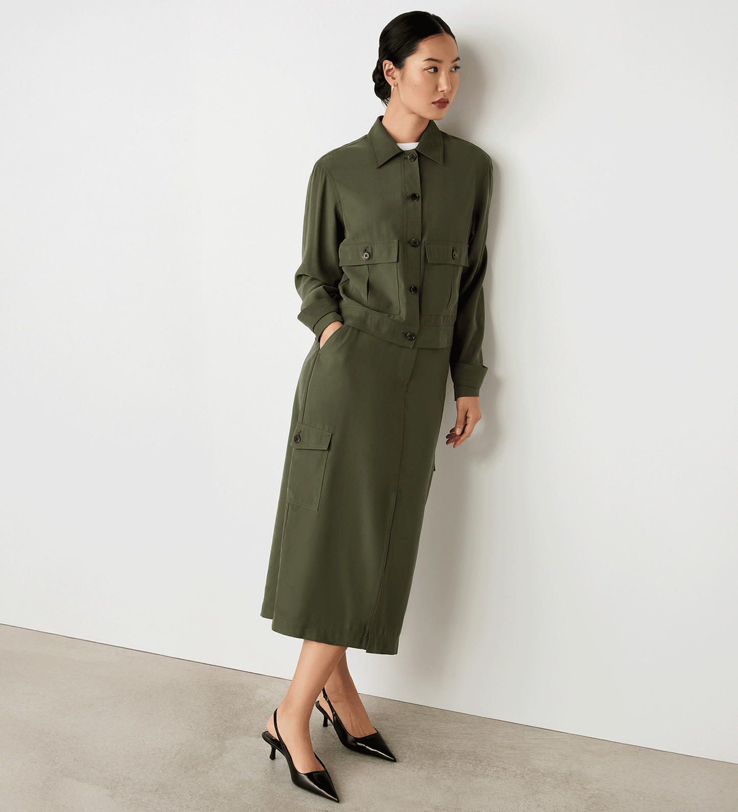 Full body view of a model wearing the Alexa Khaki Utility Jacket buttoned up facing side-on, leaning against the wall.